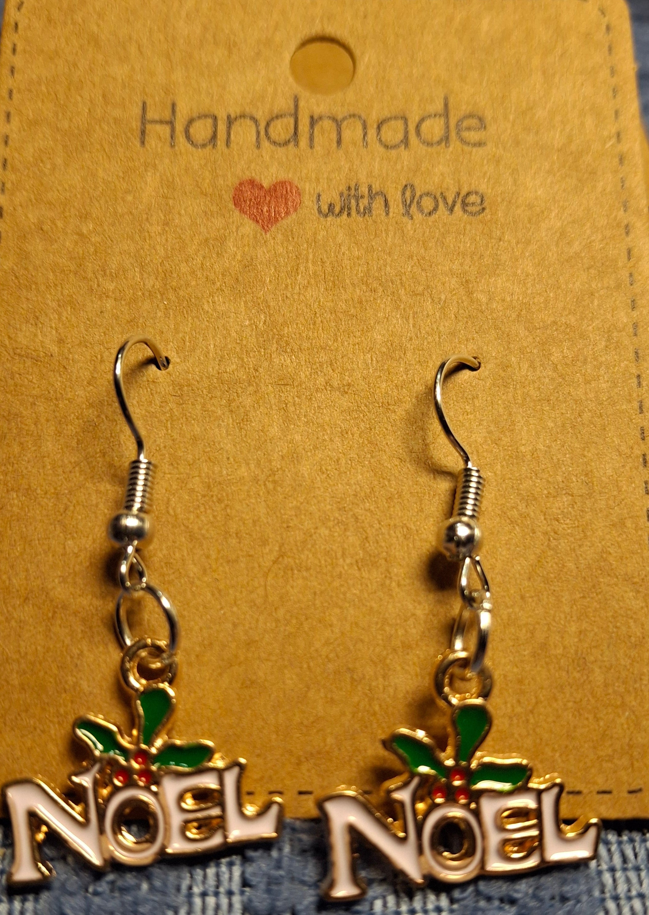 Gold 'NOEL' earrings with green holly leaves on a brown card with 'Handmade with love' text.